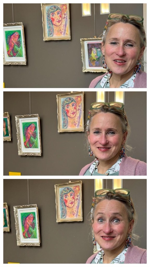 Femme souriante dans une galerie d'art, avec des peintures colorées accrochées au mur derrière elle.
